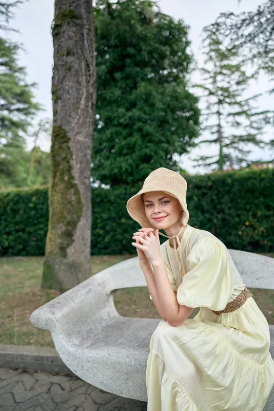 Woman in park sitting on bench wearing straw hat and flowing dress, portrait showing authenticity inclusivity, smile relaxed summer style