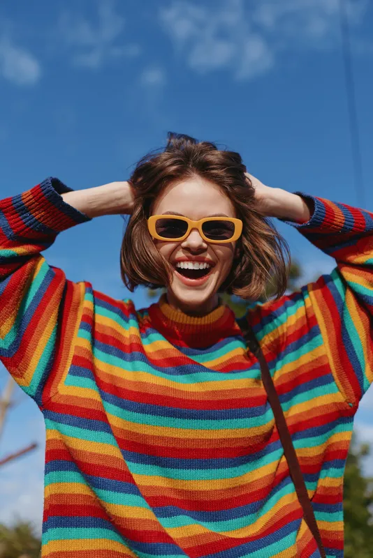 Woman, smiling, striped sweater, sunglasses, short brown hair, outdoor, blue sky, joyful, 20s model in colorful knit pullover laughing with hands in hair, casual fashion lifestyle travel vibe