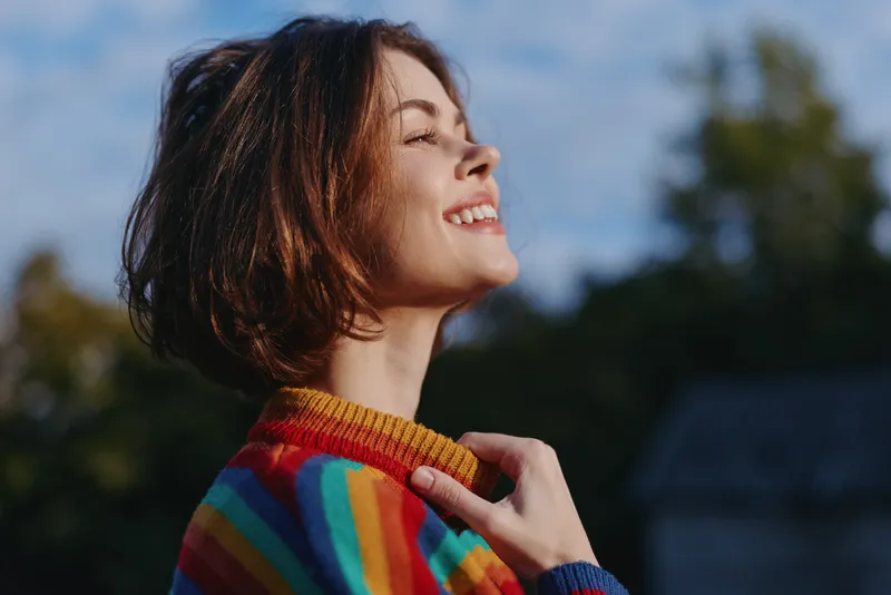 woman smile portrait outdoors in sunlight wearing rainbow sweater, happy short hair profile with closed eyes enjoying nature and warm light, relaxed youthful expression and cozy mood