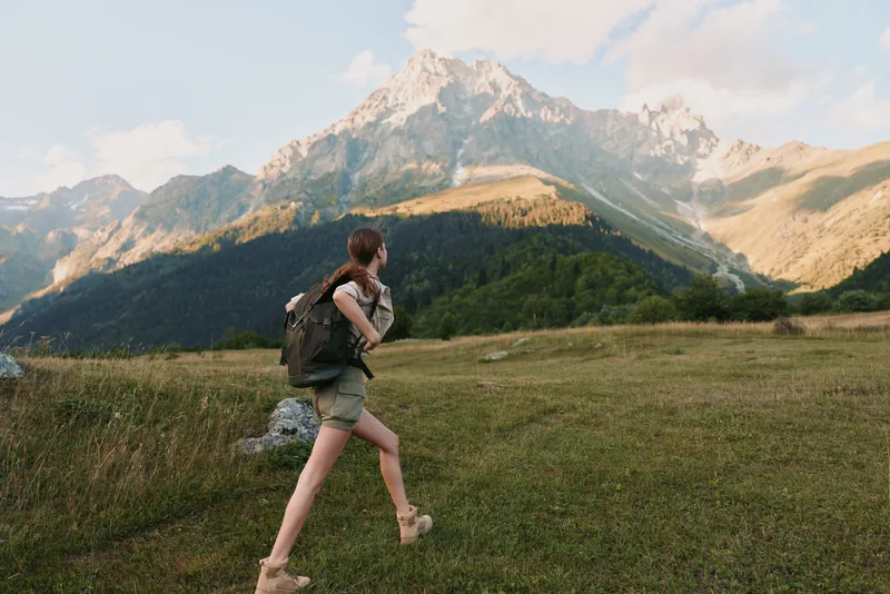 A woman with a backpack hikes through a grassy meadow towards tall mountains under a partly cloudy sky. The scene captures outdoor adventure and lifestyle in nature.