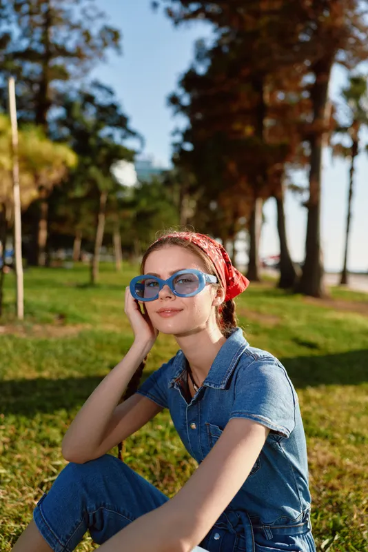 smiling woman wearing blue sunglasses and red headscarf relaxing outdoors on sunny day in green park with trees and clear sky background