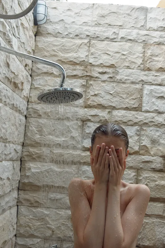 Woman covering face under outdoor shower with stone wall background, fresh water drops, natural light, and serene atmosphere, morning routine concept.