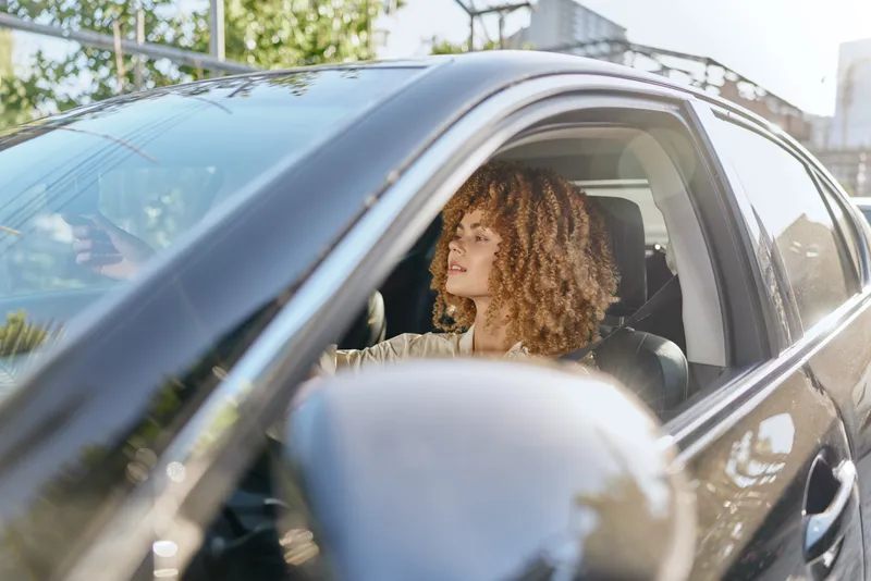 curly woman driving car on sunny day, looking thoughtful and relaxed, natural light on her face, casual outfit, modern vehicle interior