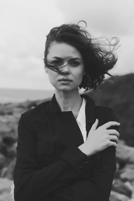 Woman on a windy coast in monochrome, wearing a jacket, hair blown, moody atmosphere, fashion photography with ocean breeze and dramatic pose