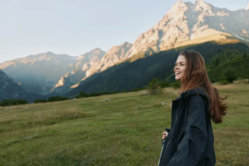 A smiling woman stands in a grassy meadow with towering mountains in the background, wearing a dark jacket. She appears relaxed and curious, enjoying fresh air in a tranquil outdoor scene.