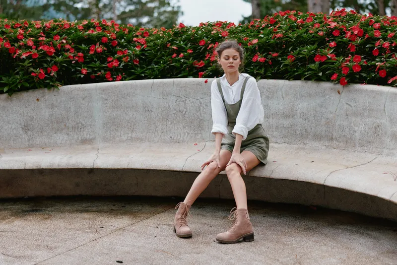 Elegant young woman in stylish outfit sitting on a bench surrounded by vibrant flowers, expressing calmness and tranquility in a natural setting