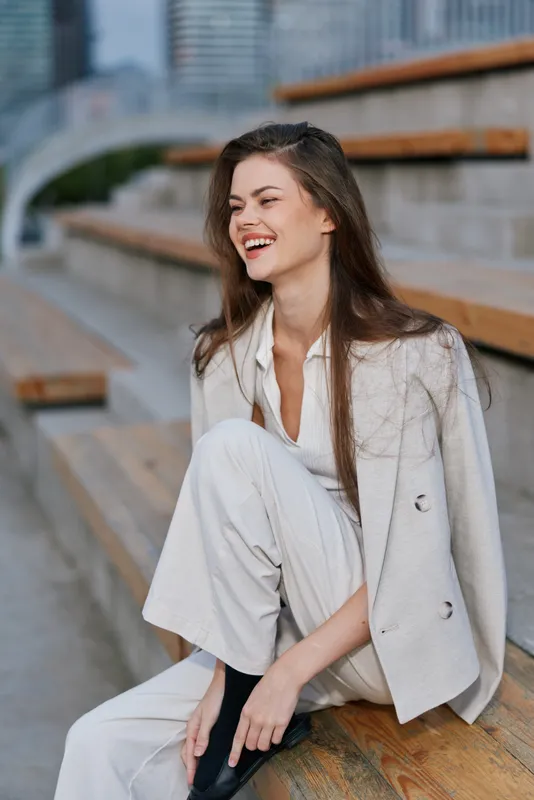 Young woman in stylish light outfit laughing outdoors on urban wooden stairs, casual fashion wear, natural emotion, spring day, modern city background
