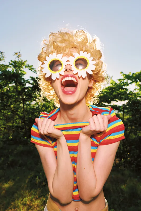 Playful woman outdoors wears a striped shirt and flower sunglasses, laughing loudly, creating a vibrant retro summer mood against a bright green backdrop in warm sunlight and joy