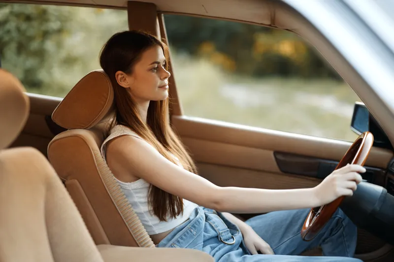 Young woman with long hair driving a vintage car, wearing a casual outfit, exuding confidence and calmness, set in a natural outdoor background.