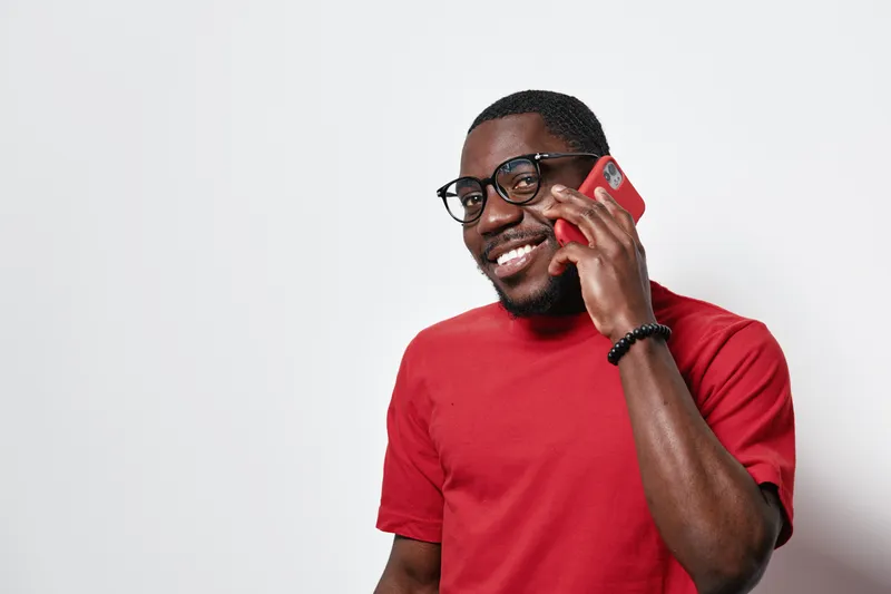 African American man with dark skin wearing glasses and red shirt smiling while talking on smartphone against white background. Casual, communication and technology concept.