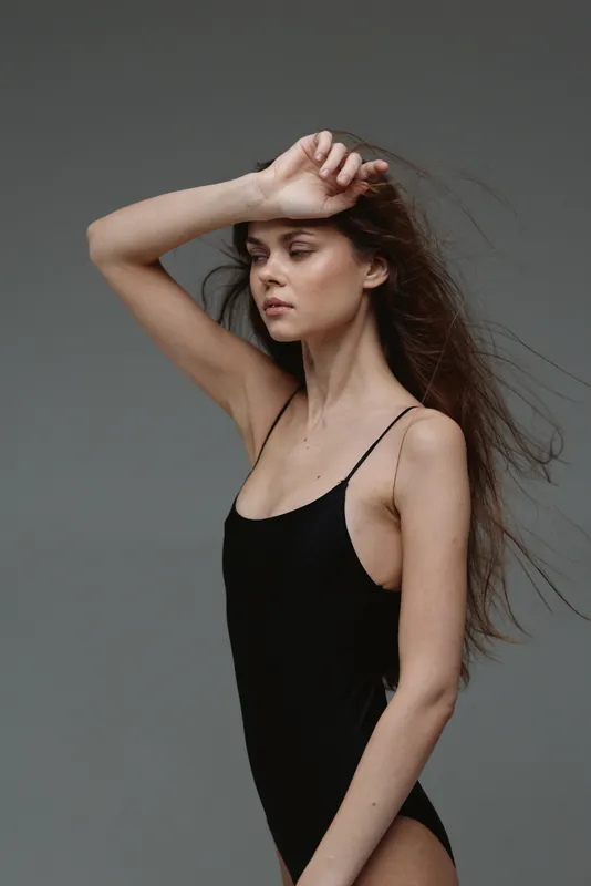 slender woman in black bodysuit posing with raised arm against gray background, studio portrait, contemplative mood