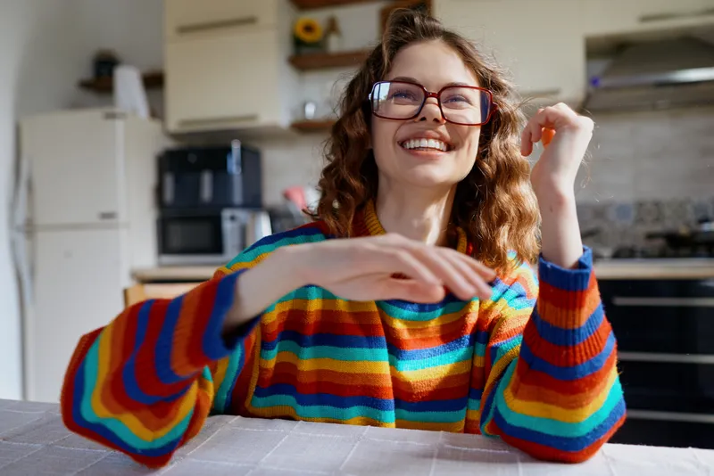 Cheerful woman indoors, wearing a colorful striped sweater, smiling at the camera, casual lifestyle moment in a bright kitchen, warm light, relaxed pose, candid and natural