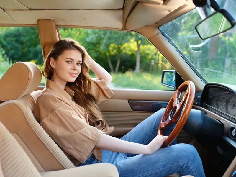 Young woman driving vintage car in nature, relaxed expression, stylish casual outfit, showcasing joy of freedom and adventure outdoors.