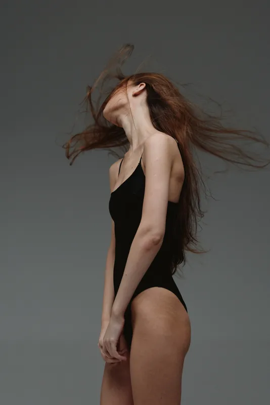 Young woman in black bodysuit with flowing hair posing against gray background, graceful movement, studio fashion portrait