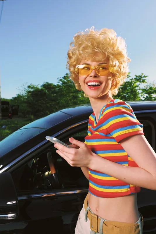 A cheerful woman with retro blond curls and yellow sunglasses stands beside a sleek car in bright sunlight. Colorful striped top highlights a playful, carefree lifestyle.