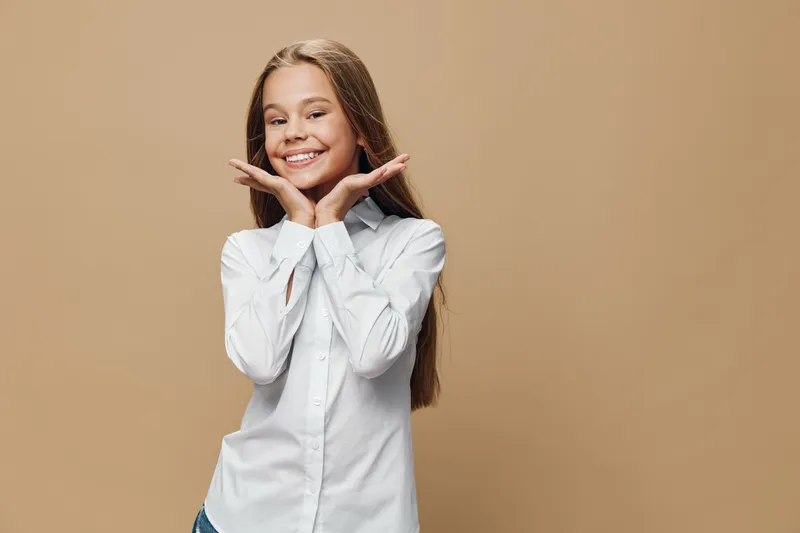 Girl smiling with long hair and white shirt posing with hands near face in studio with beige background. Child model, happiness and youth with natural expression and casual style.