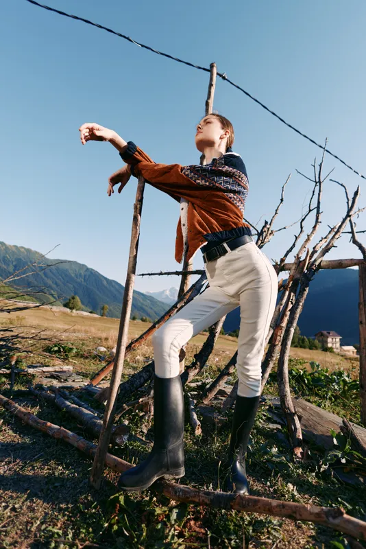 Woman outdoors in mountains posing by rustic wooden fence wearing fashionable sweater, highwaist trousers and boots, editorial pose in natural light with scenic rural landscape and sky