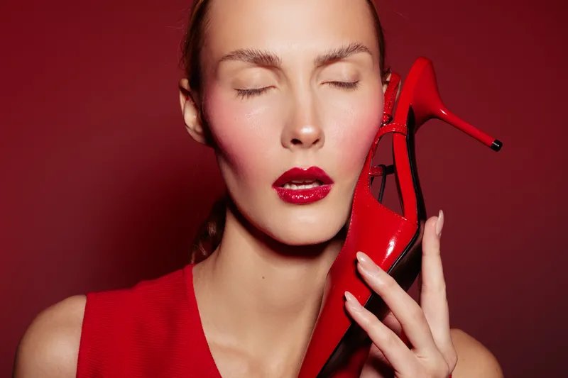 Close-up of a woman with closed eyes holding a red high heel shoe near her face. Red background and red outfit create a bold fashion and beauty concept with glossy lips.