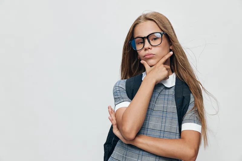 Girl with long hair and glasses wearing a dress with backpack thinking and posing with hand on chin on white background. Youth portrait with casual style and serious expression.