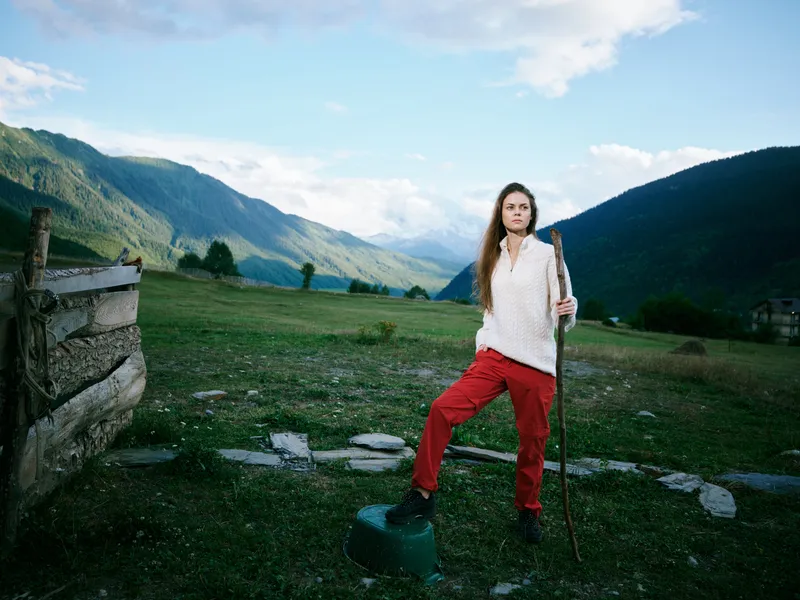 Woman standing with a walking stick in a wide alpine meadow, mountains in the background, clear sky, fence on the left, rugged terrain, casual outdoor gear, calm countryside scene, exploring nature