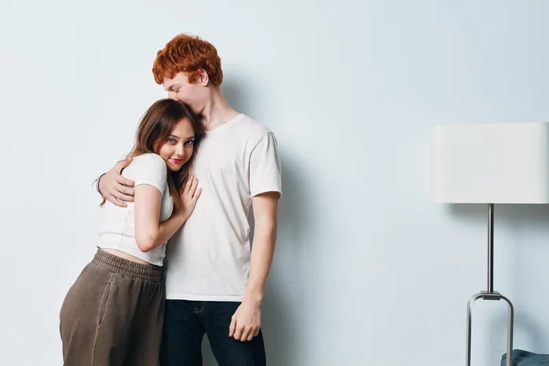 Young couple embracing tenderly near white wall in casual clothes indoors, showing affection and closeness in cozy home environment with soft lighting and relaxed mood.