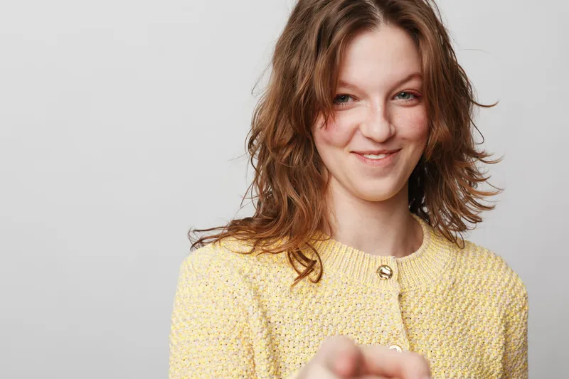Smiling young woman with wavy brown hair wearing yellow knitted sweater points forward. Studio portrait on light grey background. Concept of confidence, friendliness, and positive attitude.