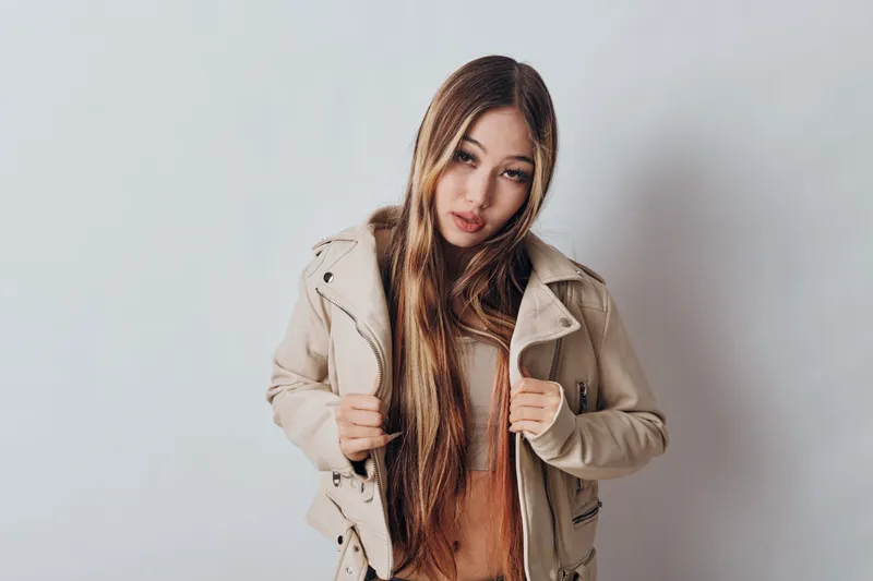 Young woman standing against white wall wearing beige jacket and sweater with long hair and casual pose for fashion portrait and style in studio setting.