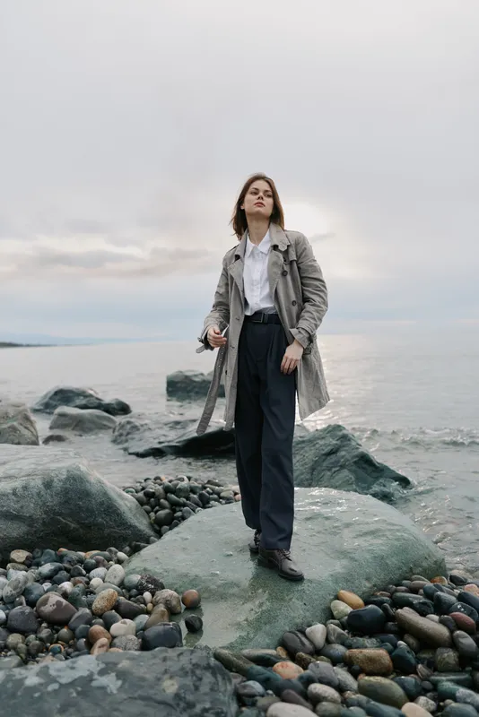 Stylish woman standing on rocky beach wearing trench coat and dark pants. Overcast sky and calm sea create moody atmosphere. Concept of fashion, confidence, and outdoor lifestyle.