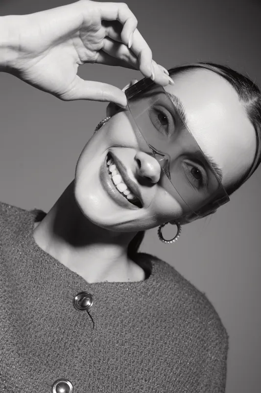Happy Caucasian woman with light skin wearing oversized transparent glasses and textured sweater smiles at camera in black and white studio portrait.