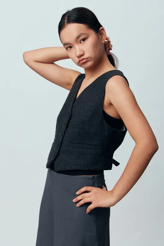 Young woman posing with hand on hip and the other behind her head wearing a sleeveless vest and pants against a plain light background with a confident expression.