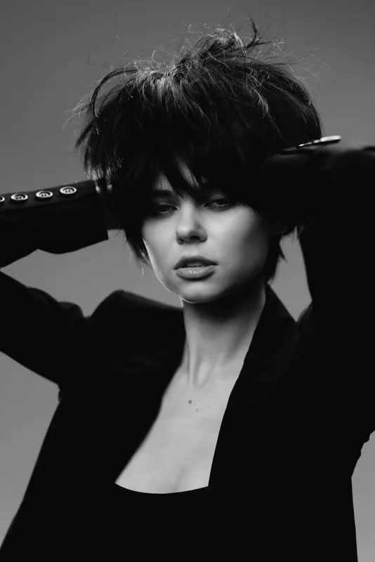 Black and white portrait of a stylish woman with short hair and intense expression wearing elegant dark clothing.