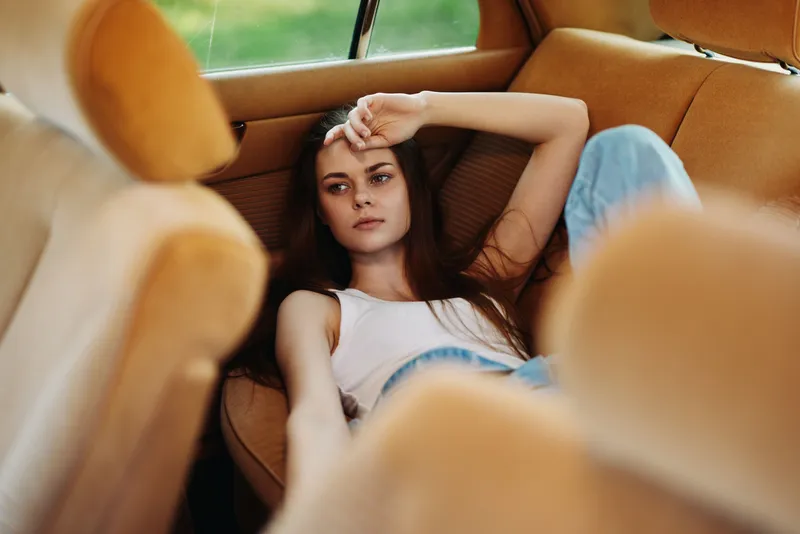 Young woman relaxing on brown car seats, wearing casual attire, reflecting deep thoughts and emotions in a serene, natural setting with soft lighting.
