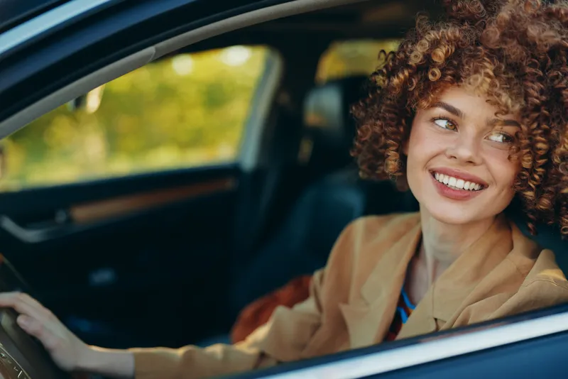 Joyful woman driver with curly hair sits in a car and smiles, a candid lifestyle moment with warm retro film color and everyday travel vibe, bright and relaxed.