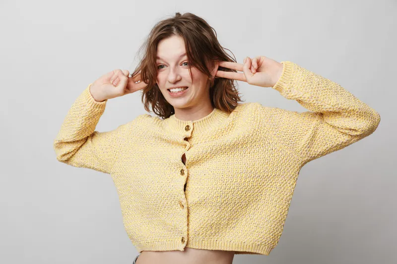 Young woman in yellow knitted sweater makes playful face and covers ears with fingers. Studio portrait on grey background. Concept of fun, emotions, youth, and casual fashion.