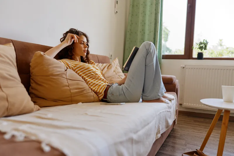 Young woman lounges on a cozy sofa at home, wearing a striped shirt and jeans, enjoying a calm moment in a warm living room, soft light creating a relaxed mood