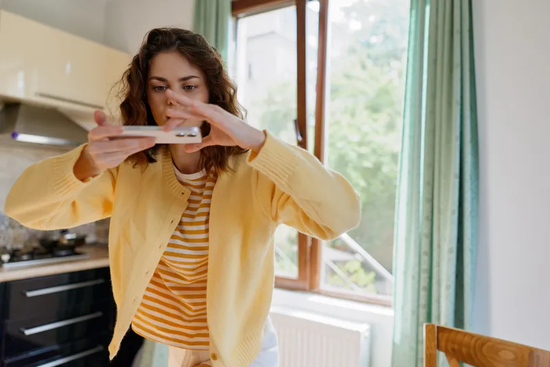 Casual woman home lifestyle scene, she holds a smartphone, taking a photo in a bright kitchen, wearing a yellow cardigan and striped shirt, cheerful, candid moment.