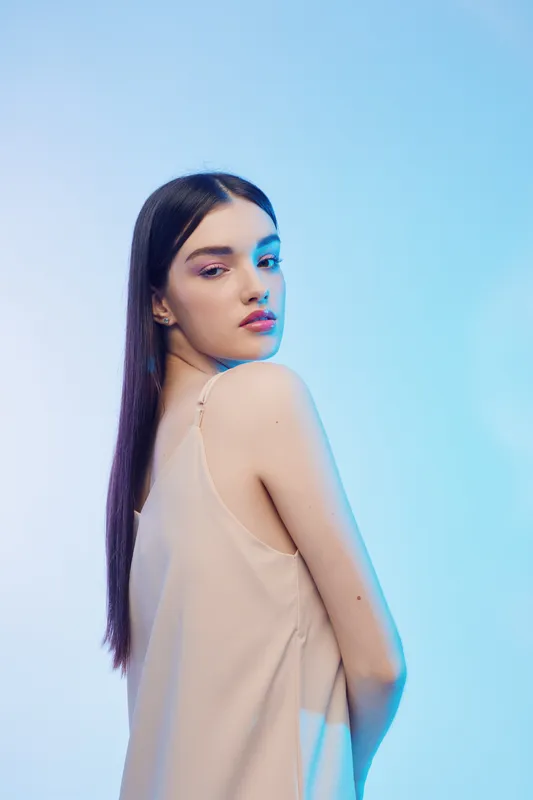Young woman with long dark hair poses in a beige sleeveless top against a blue background. The image highlights beauty, fashion, and elegance with soft lighting. Concept for style.