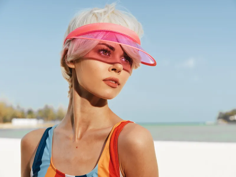 Portrait of a stylish woman at the beach wearing a colorful swimsuit and a translucent pink visor, sunlit skin, confident gaze, and breezy seaside ambience.