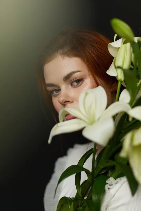 A serene young woman with red hair partially obscured by white lilies, conveying a sense of calm and beauty in a dark background, perfect for exploring themes of nature and femininity