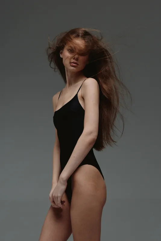Young woman with long flowing hair in black bodysuit posed against gray background