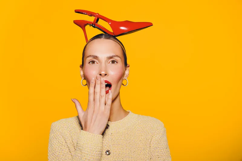 Surprised woman with red high heel shoes on her head covers mouth with hand. Bright yellow background and beige sweater create playful and quirky fashion concept with unexpected style.