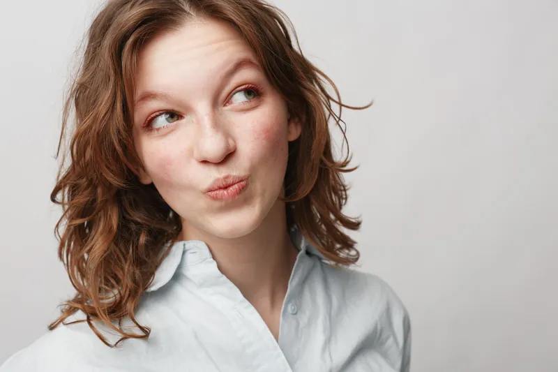 Young woman with curly hair wearing white shirt looks sideways with playful expression on light background. Concept of casual style, emotions, and natural beauty portrait.