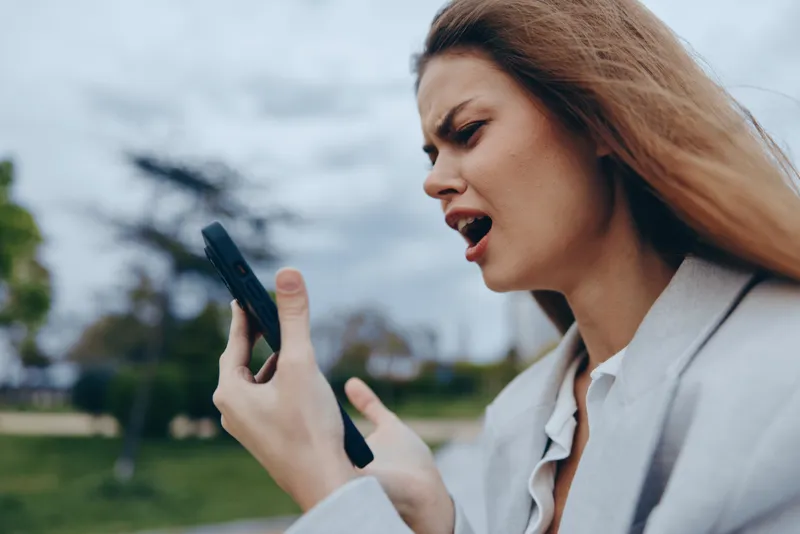 frustrated woman holding smartphone outdoors with intense emotion in casual business attire expressing financial crisis and stress in overcast urban park