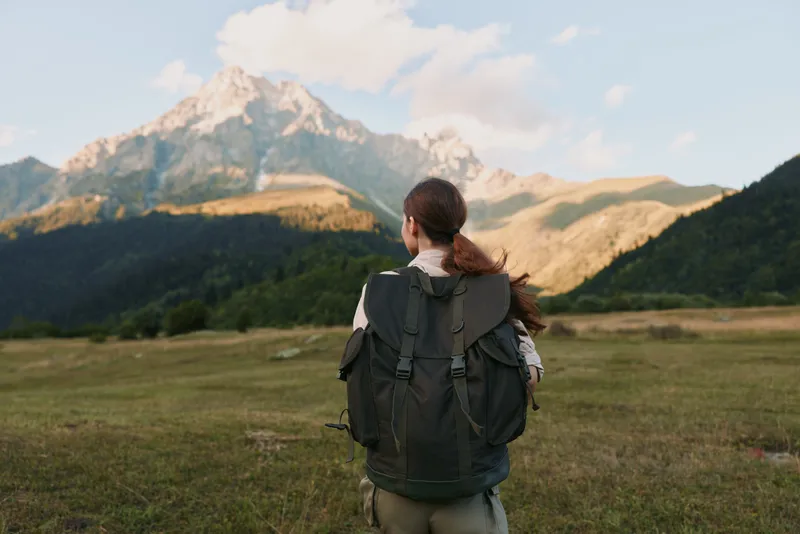 A young woman with a backpack stands in a wide alpine meadow, facing distant snow capped peaks, conveying solitude, exploration, and a hopeful sense of adventure.