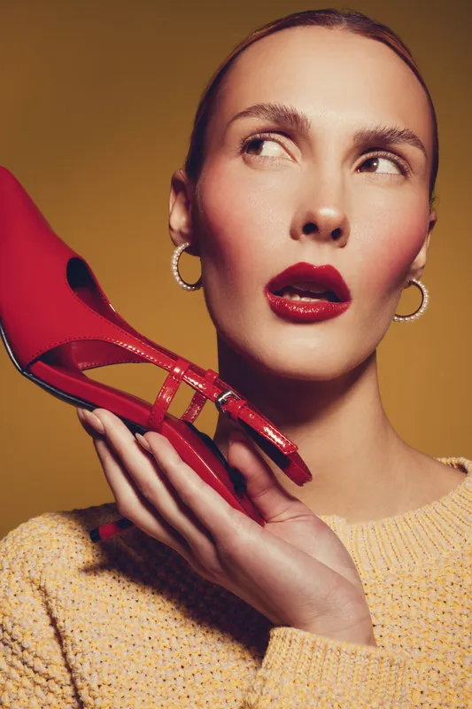 Close-up of a woman with red lipstick holding a red high heel shoe near her face. Fashion concept with bold makeup, hoop earrings, and a yellow textured sweater on warm background.