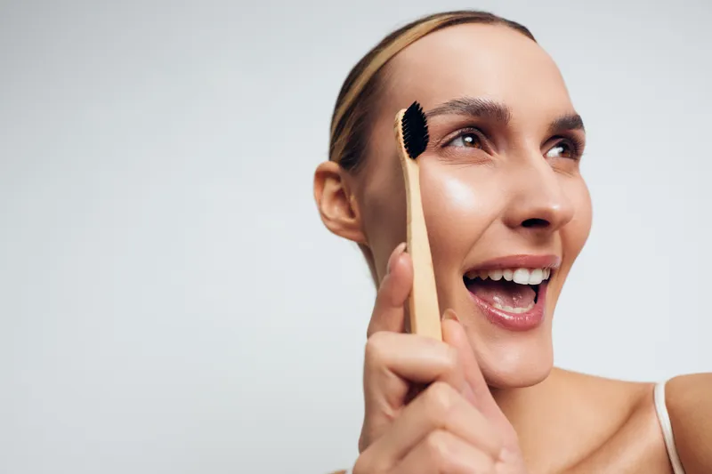 Young woman smiling with open mouth while applying mascara on eyelashes, close up portrait, natural makeup, bright background, beauty routine moment