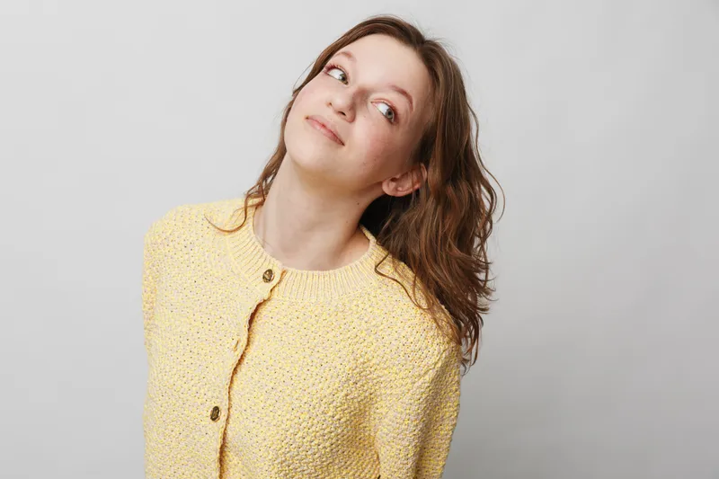 Young woman with brown hair wearing yellow knitted cardigan looks up with a thoughtful expression on a plain light background. Concept of contemplation, casual style, and natural beauty.
