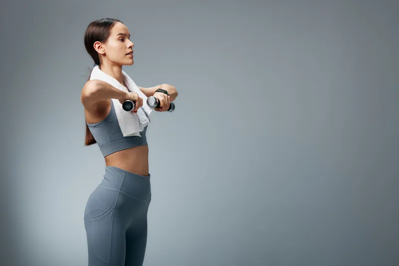 Fit young woman in grey activewear performs an upright row with small dumbbells, towel draped around her neck, focused side profile in a minimalist studio with neutral grey background.