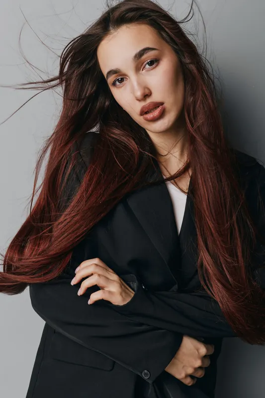 Young woman with long hair and natural makeup posing in black blazer against gray background. Fashion model looking at camera with confident expression and casual posture indoors.