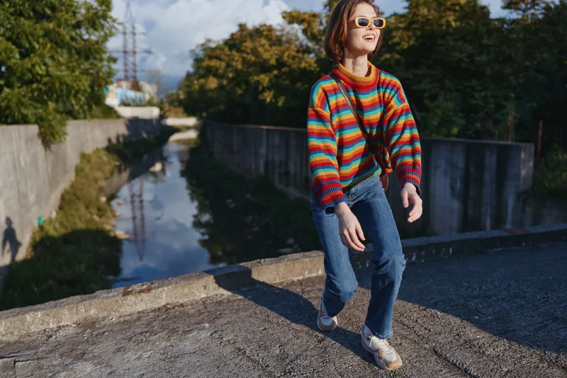 Girl in a colorful striped sweater and sunglasses walking and smiling along an urban canal in autumn, preteen model in jeans, casual outfit with beanie and sneakers, lifestyle travel mood.